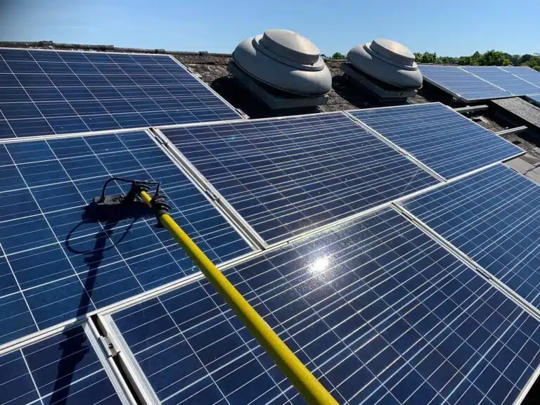 A long yellow pole with a brush cleaning solar panels on a rooftop under a clear blue sky, showcasing vibrant sunlight reflections.
