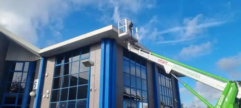 A worker on a cherry picker performs window cleaning near the top of a commercial building with blue accents.