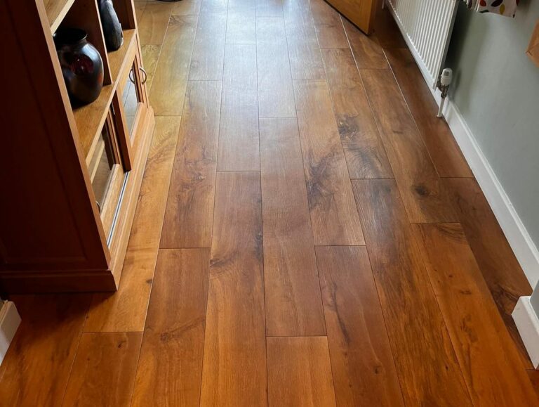 A wooden floor with rich brown tones and visible grain patterns, leading towards an open doorway and a wooden cabinet on the left.