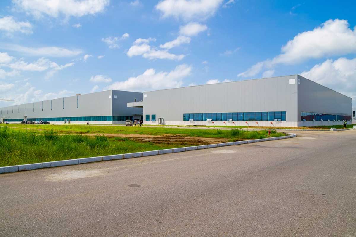 A large, modern gray warehouse building with windows under a blue sky, surrounded by green grass and a paved road.